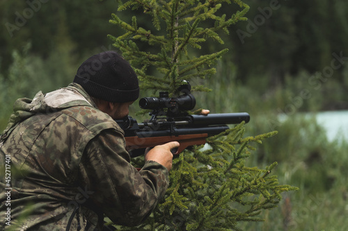The hunter aims with a rifle. A man in camouflage is preparing to shoot. Hunting in the woods with a sniper rifle