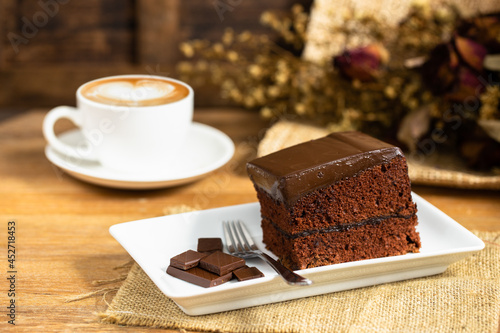 Chocolate cake decorated by pice of chocolate and fork on white plate with cup of latte coffe with heart shape latte art on wooden table