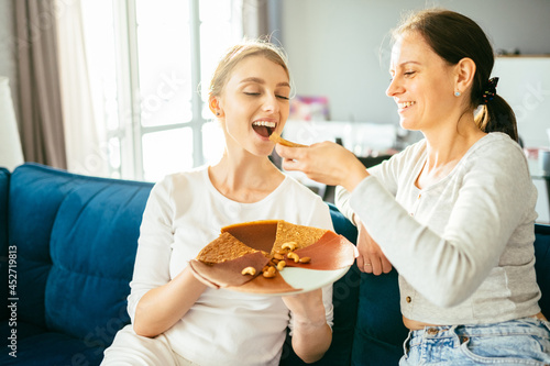 Happy cheerful young woman and her nice looking mother sitting on coach try fruit marshmallow while preparing berry pastille, dish with fresh fruits at home. Healthy food, homemade sweets concept.