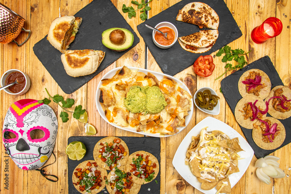 Top view image of set of Mexican dishes and catrina mask. Cochinita pibil tacos, pastor tacos, nachos with guacamole, quesadillas, jalapeños, limes and cilantro Top view image of set of Mexican dishes and catrina mask. Cochinita pibil tacos, pastor tacos, nachos with guacamole, quesadillas, jalapeños, limes and cilantro
