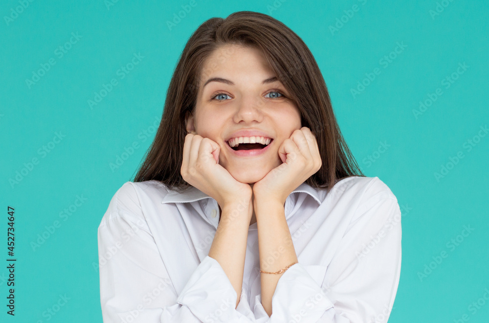 Indoor portrait of young attractive European girl isolated on blue background looking straight at camera propping up her chin with fists feeling great joy, smiling and laughing at joke Indoor portrait of young attractive European girl isolated on blue background looking straight at camera propping up her chin with fists feeling great joy, smiling and laughing at joke