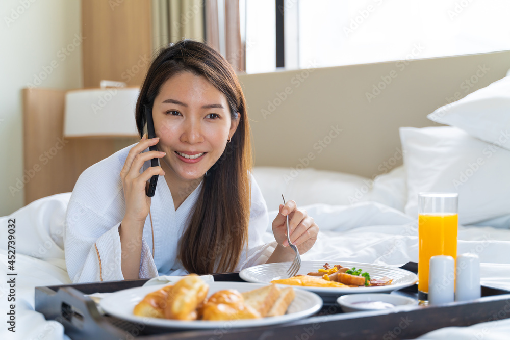 Portrait beautiful young Asian woman wearing bathrobe talking on a phone while eating breakfast on a bed in bedroom