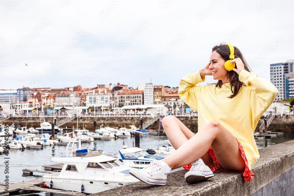 Young woman sat on promenade of port city listening to music. Boats in the background