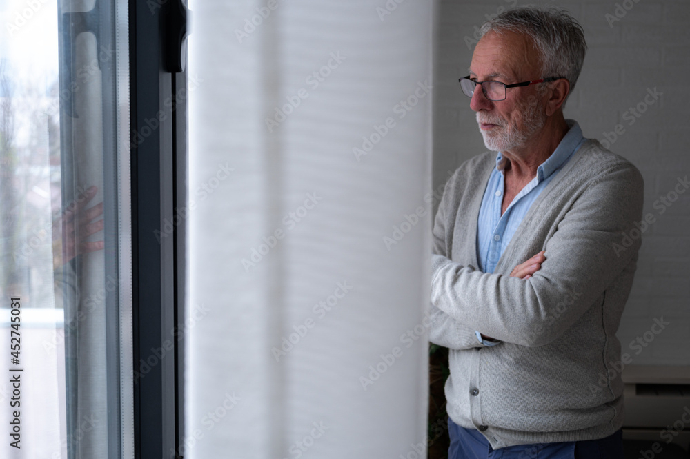 Fototapeta premium Portrait of an elderly bearded man with eyeglasses looking sad through the window of his apartment. Loneliness