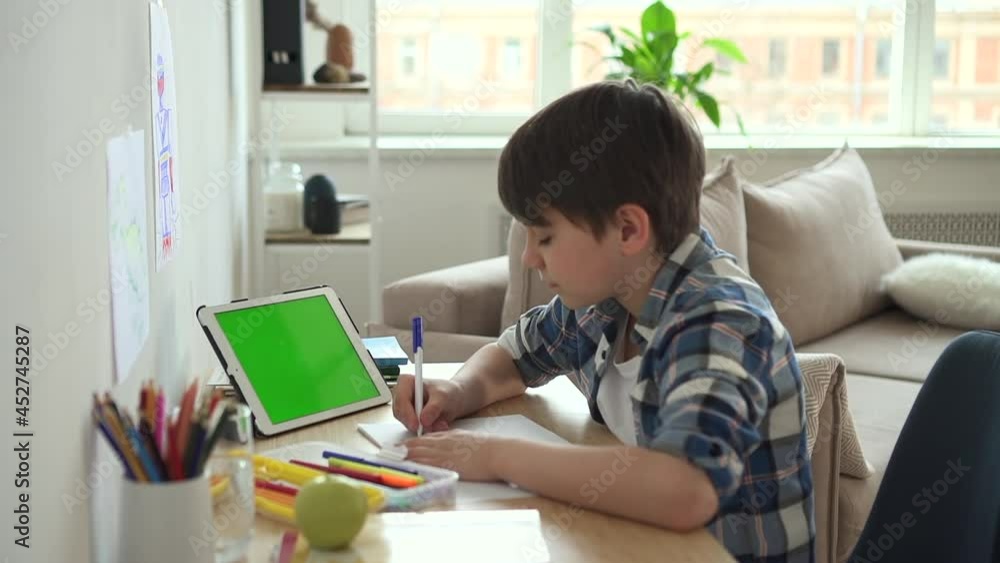 Schoolboy writing in notebook and sitting at table with tablet during online conference spbd. Closeup view of cute boy writes in notebook and looks at device screen, does study practice and sits at Schoolboy writing in notebook and sitting at table with tablet during online conference spbd. Closeup view of cute boy writes in notebook and looks at device screen, does study practice and sits at