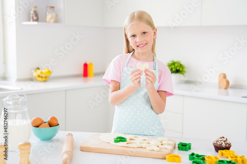 Photo of young happy joyful small girl hold hand cookie heart shape baker smile indoors inside house kitchen
