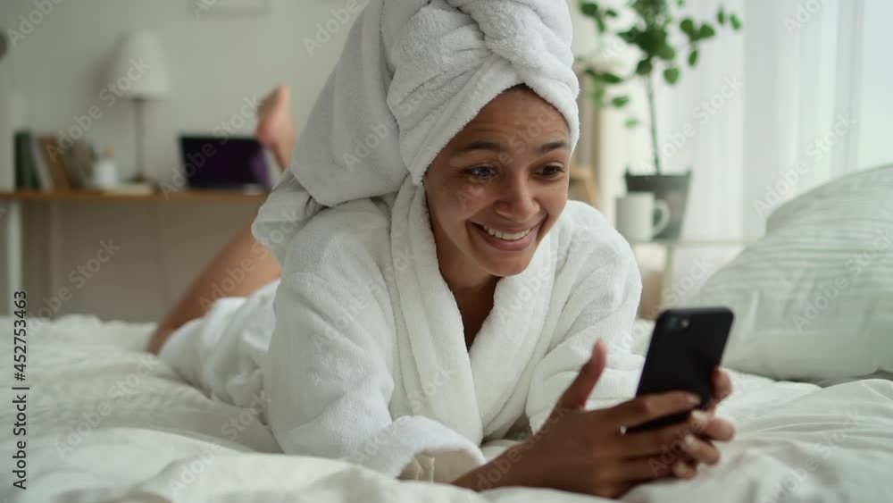 Beauty procedure undergoing at home. Spbd Surprised African-American woman with towel turban nods head left-right denying at videochat on cellphone on bed in room Beauty procedure undergoing at home. Spbd Surprised African-American woman with towel turban nods head left-right denying at videochat on cellphone on bed in room