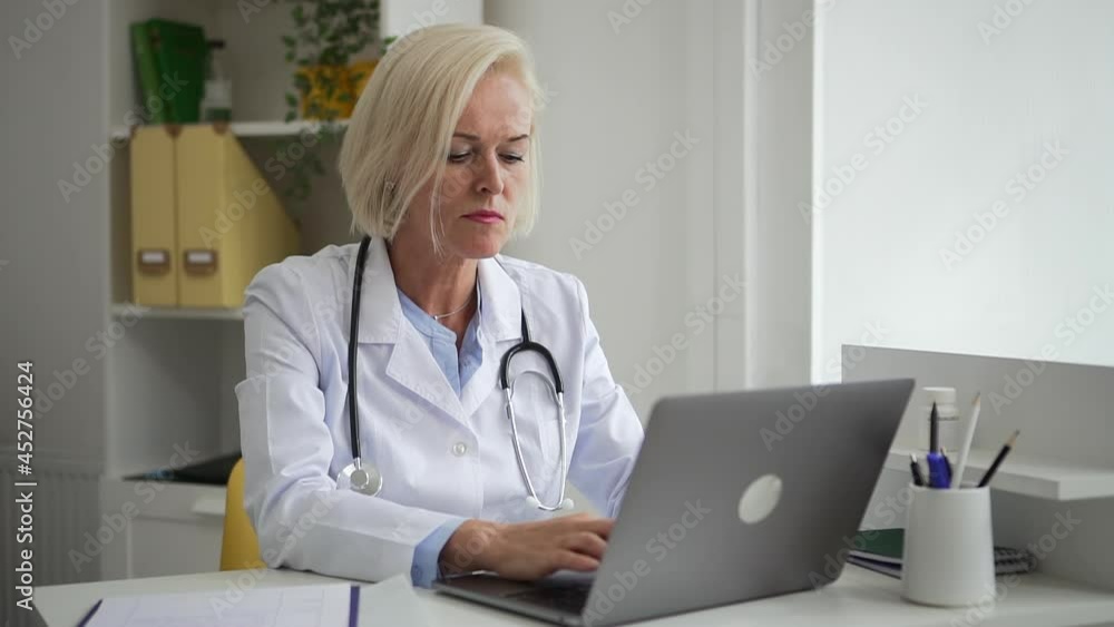 Doctor telehealth remote service. Spbd Concentrated middle aged lady therapist in coat types on laptop keyboard looking at papers in hospital office Doctor telehealth remote service. Spbd Concentrated middle aged lady therapist in coat types on laptop keyboard looking at papers in hospital office