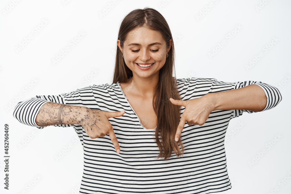 Beautiful woman pointing and looking down with pleased smile, standing in casual striped blouse against white background Beautiful woman pointing and looking down with pleased smile, standing in casual striped blouse against white background