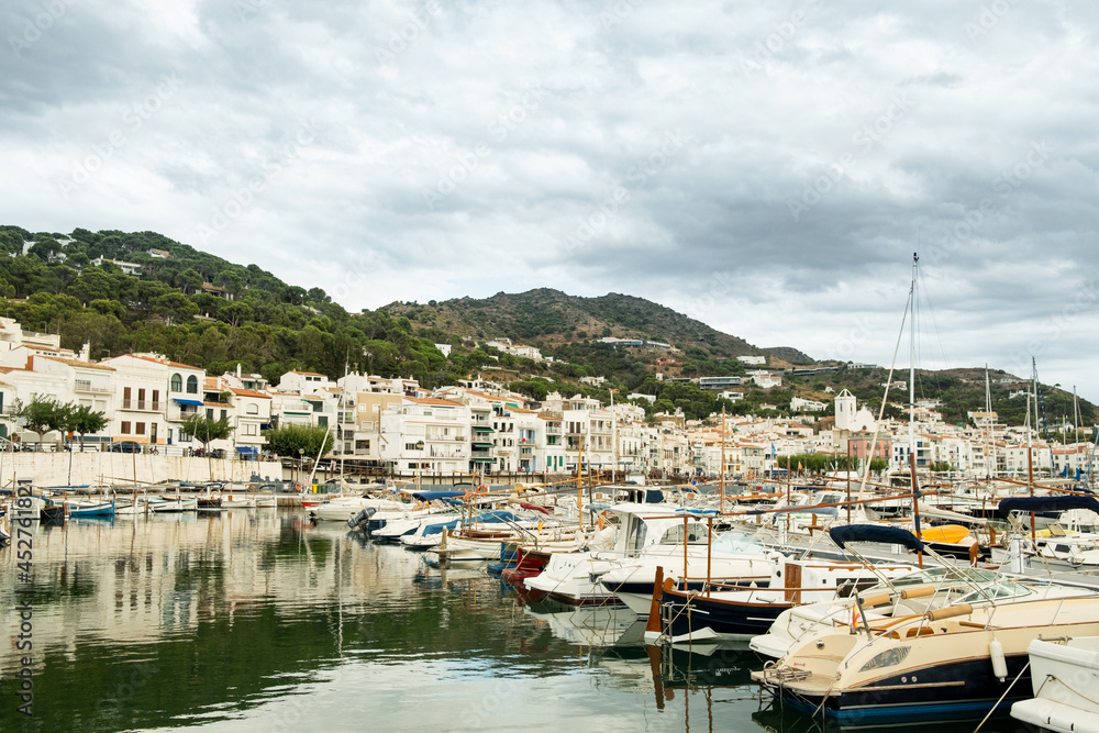 View of small spanish town of Port de la Selva harbor in the Costa Brava in Catalonia, with small white fishing boats on a cloudy summer day during tourist season.