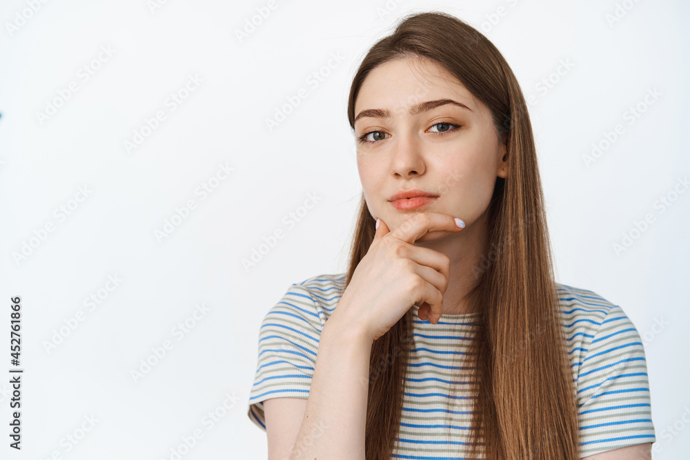 Thoughtful girl looking at camera with hand on chin, thinking, listening to smth interesting, white background Thoughtful girl looking at camera with hand on chin, thinking, listening to smth interesting, white background