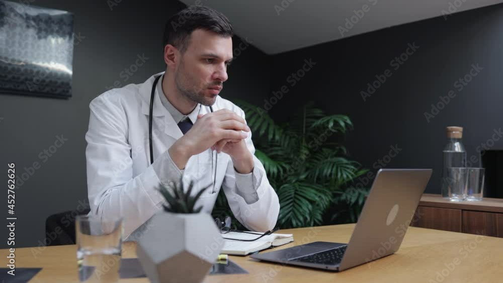 Male doctor using laptop and talking during online conference at table in modern clinic spbi. Young Caucasian man looks at computer screen and speaks something, sits at desk in light interior. Closeup Male doctor using laptop and talking during online conference at table in modern clinic spbi. Young Caucasian man looks at computer screen and speaks something, sits at desk in light interior. Closeup