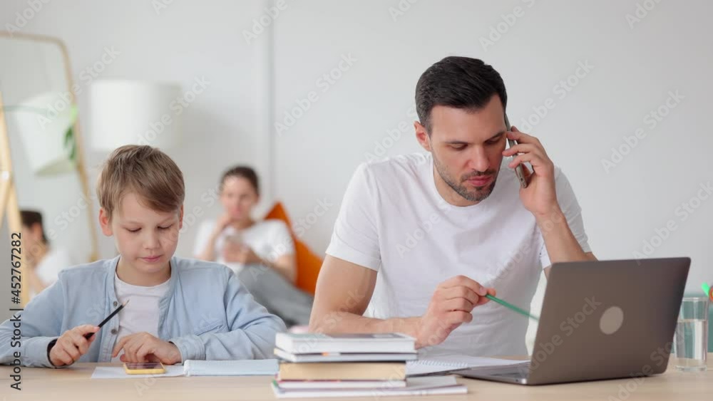 Dad talking on phone and son playing with gadget while sitting at table in home room spbi. Front view of young man has mobile conversation and works with laptop, cute child plays with smartphone and Dad talking on phone and son playing with gadget while sitting at table in home room spbi. Front view of young man has mobile conversation and works with laptop, cute child plays with smartphone and