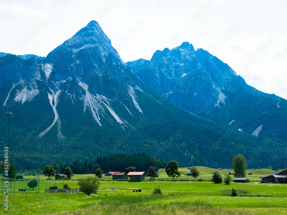 Obraz premium Panoramic view from Grubigstein, with rugged Zugspitze in background under blue sunny sky with cumulus clouds. Green valley at the foot of majestic alpine mountains, in Lermoos, Tyrol
