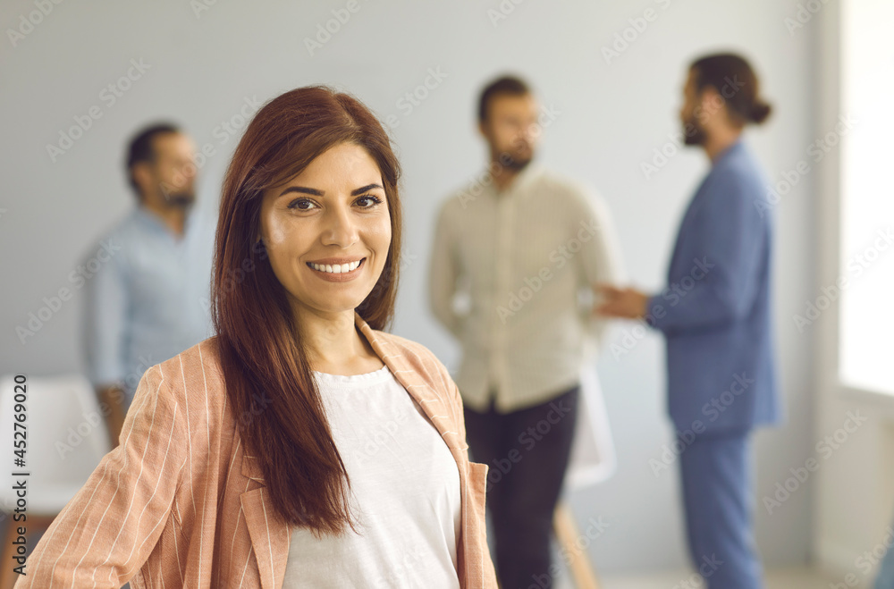 Close up portrait of a successful young business woman looking at camera with confident expression. Smiling woman on a background of people talking to each other in a bright room. Confidence concept. Close up portrait of a successful young business woman looking at camera with confident expression. Smiling woman on a background of people talking to each other in a bright room. Confidence concept.