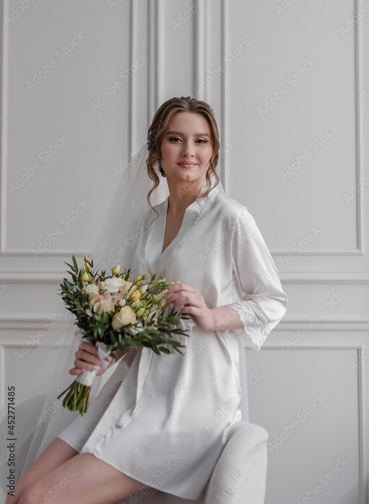 A beautiful young girl in a dressing gown at the window with a bouquet in her hands is a blonde with hair and makeup in a white room. A beautiful young girl in a dressing gown at the window with a bouquet in her hands is a blonde with hair and makeup in a white room.