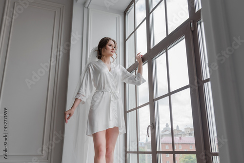 A beautiful young girl in a white robe and a veil on a chair in a white room with hair and makeup, a smile, happiness.