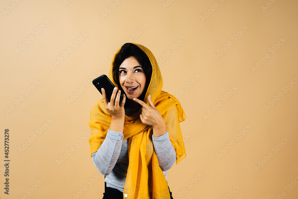 Obraz premium Smiling young arabian muslim woman in hijab light clothes posing isolated on yellow background. People religious Islam lifestyle concept. copy space. Hold mobile phone sending audio message