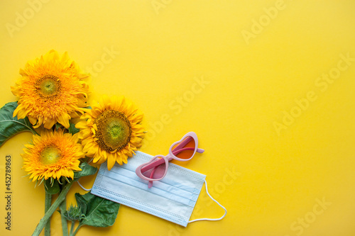 Arrangement of pink heart-shaped sunflower-shaped sunglasses and protective medical mask on bright yellow background. High quality photo