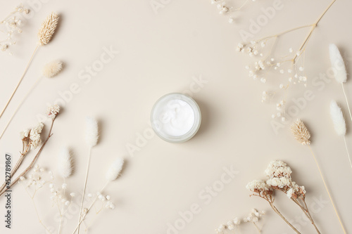 Jar of cosmetic cream with flowers on pastel beige background. Flat lay
