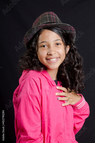  Curly hair latin girl wearing a hat smiles with hand on chest, black background 