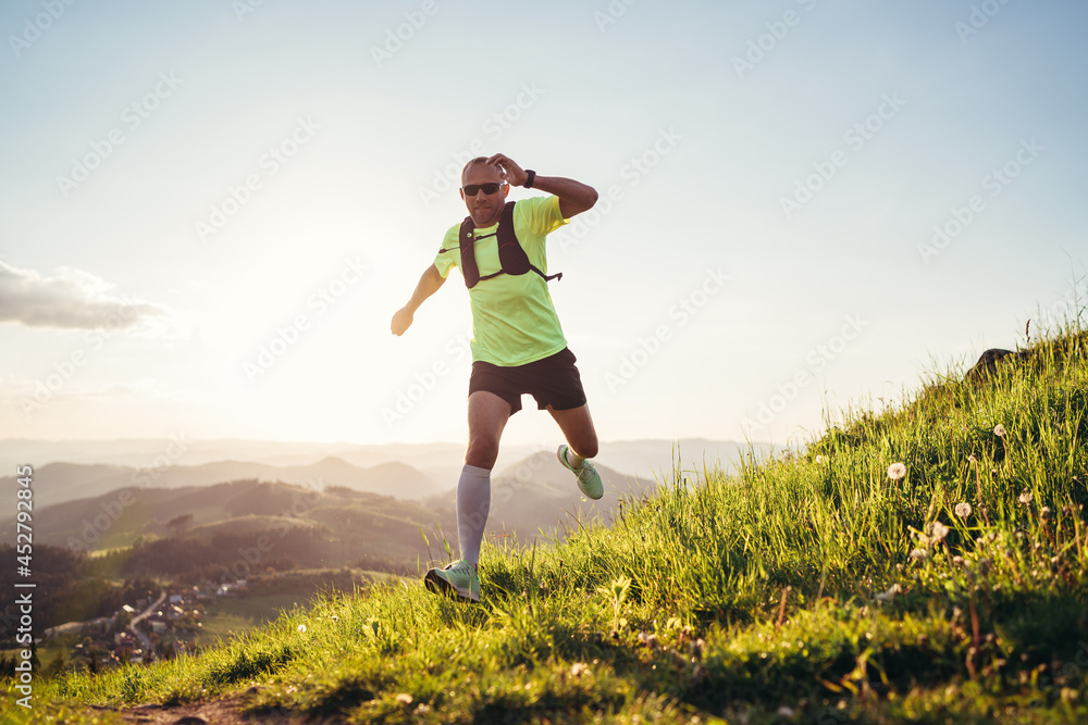 Active mountain trail runner dressed bright t-shirt with a backpack in sports sunglasses running endurance ultramarathon race by picturesque hills at sunset time. Sporty active people concept image.. Active mountain trail runner dressed bright t-shirt with a backpack in sports sunglasses running endurance ultramarathon race by picturesque hills at sunset time. Sporty active people concept image..