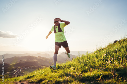 Active mountain trail runner dressed bright t-shirt with a backpack in sports sunglasses running endurance ultramarathon race by picturesque hills at sunset time. Sporty active people concept image..