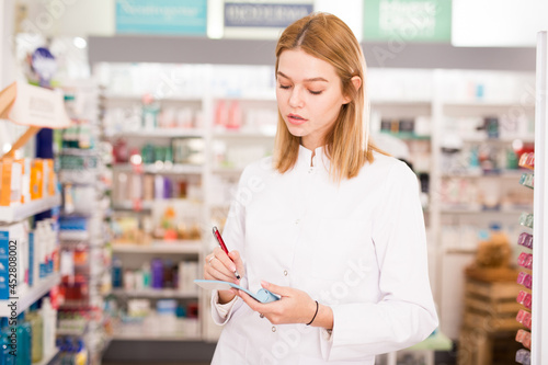 Smiling female pharmacist checking assortment of drugs in pharmacy