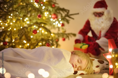 Little boy waiting Santa Claus under tree on Christmas Eve. Child is sleeping. Santa Claus brought gifts. Magic at Christmas and New Year night.