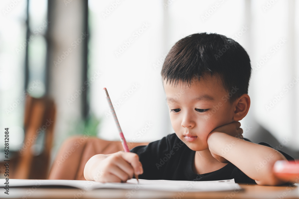 Asian Boy Doing homework with the intention. Child boy holding pencil writing, A boy drawing on white paper at the table, Elementary school and home schooling, Distance Education concept.. Asian Boy Doing homework with the intention. Child boy holding pencil writing, A boy drawing on white paper at the table, Elementary school and home schooling, Distance Education concept..