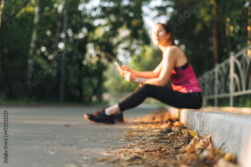 Blurred photo of Young slim woman brunette in sport clothes resting and using mobile phone at autumn park on golden hour sunrise time. Health and wellness, fitness lifestyle