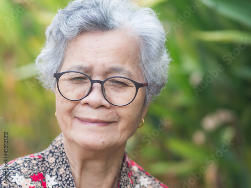Portrait of a senior woman with short gray hair, wearing glasses, smiling and looking at the camera while standing in a garden. Aged people and relaxation concept