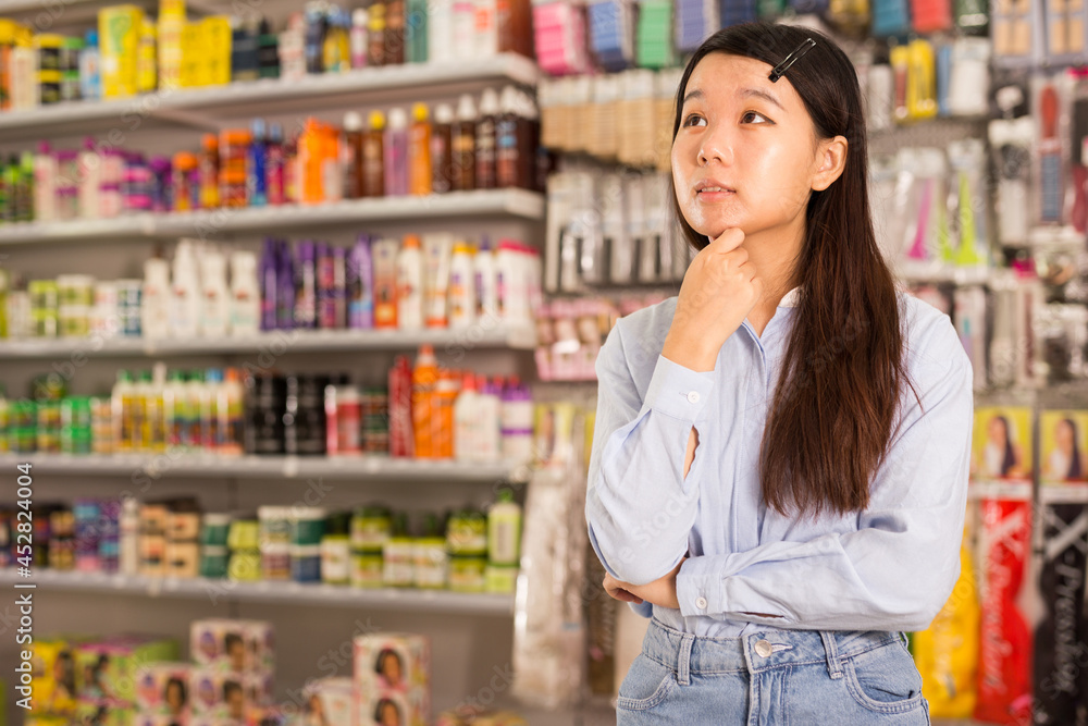 Portrait of puzzled Asian girl having doubts about choice of hair care products in special store