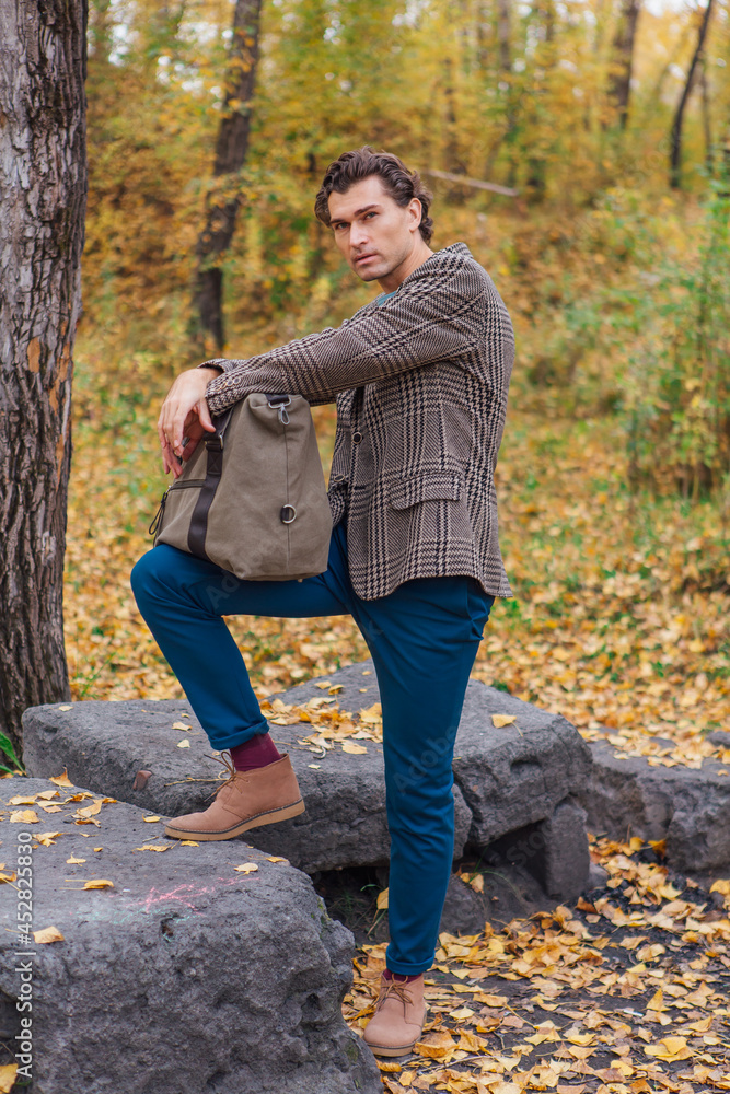 Tall handsome man with a bag standing near the rocks in the autumn forest Tall handsome man with a bag standing near the rocks in the autumn forest