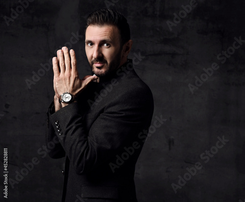 Portrait of bearded mature businessman in jacket with mandarin collar standing sideways with hands put together, looking at camera, thinking over dark background with copy space. Side view