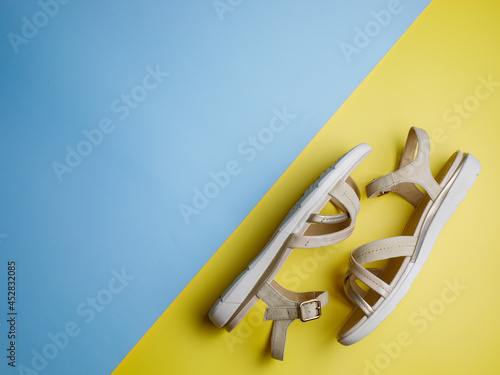 Stylish beige leather summer comfortable women's sandals on bright yellow and blue paper background. Concept of recreation by sea and summer vacation. Flat lay, top view, copy space. photo