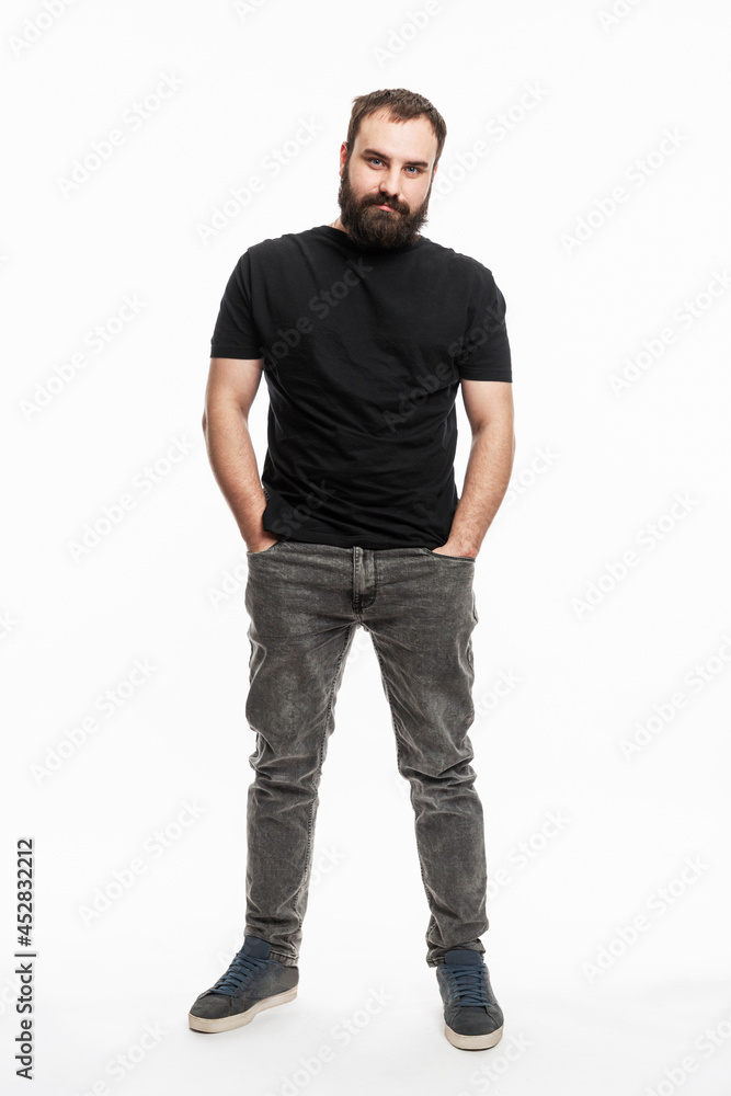 A man with a beard is standing with his hands in his pockets. Young man in jeans and a black T-shirt. Full height. White background. Vertical. A man with a beard is standing with his hands in his pockets. Young man in jeans and a black T-shirt. Full height. White background. Vertical.
