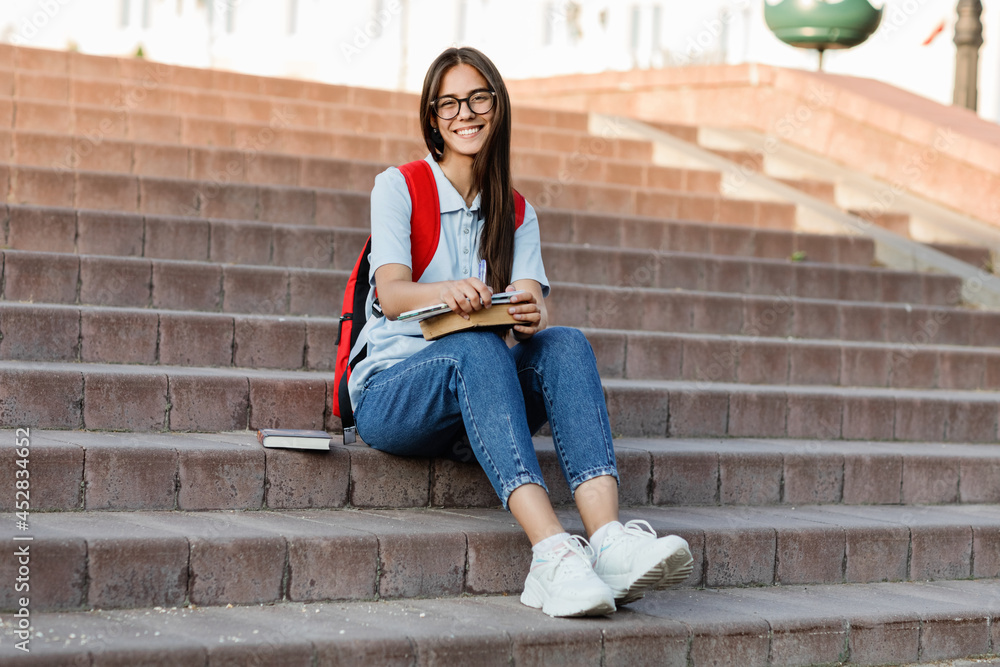 A focused student with glasses, sitting on the stairs, writes in her notebook. Preparation for a lecture or exam A focused student with glasses, sitting on the stairs, writes in her notebook. Preparation for a lecture or exam