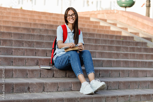 A focused student with glasses, sitting on the stairs, writes in her notebook. Preparation for a lecture or exam © Sviatlana