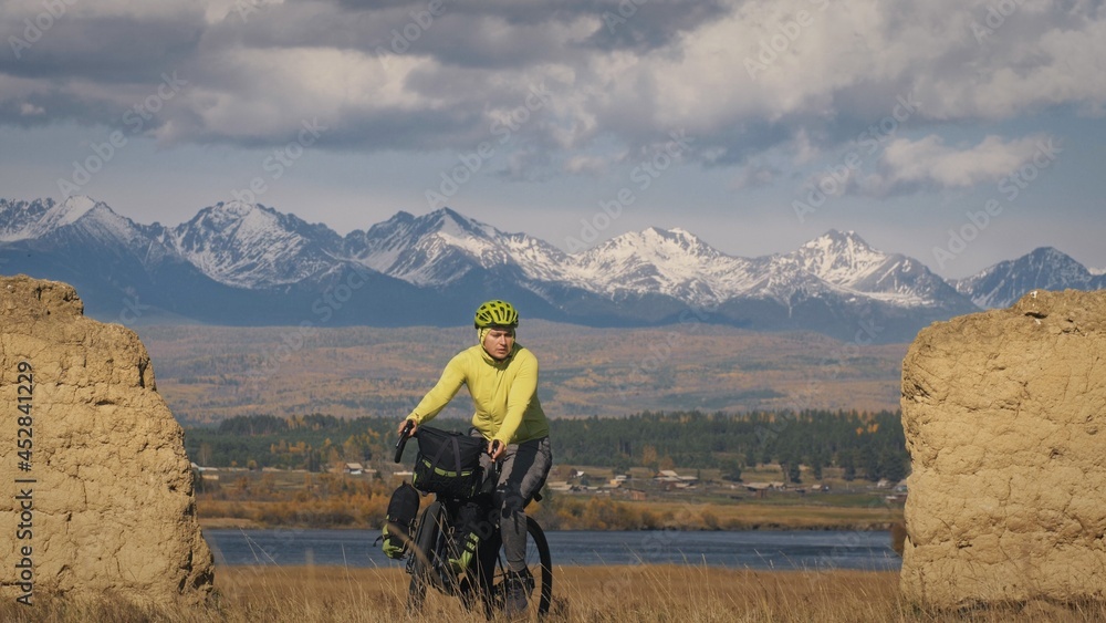 Fototapeta premium The man travel on mixed terrain cycle touring with bikepacking. The traveler journey with bicycle bags. Sport bikepacking, bike, sportswear in green black colors. Mountain snow capped, stone arch.