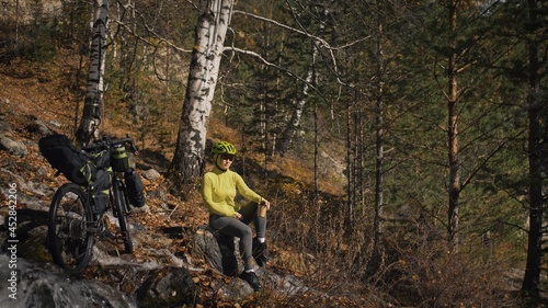 The woman travel on mixed terrain cycle touring with bikepacking. The traveler journey with bicycle bags. Sportswear in green black colors. The trip in magical autumn forest, river stream.