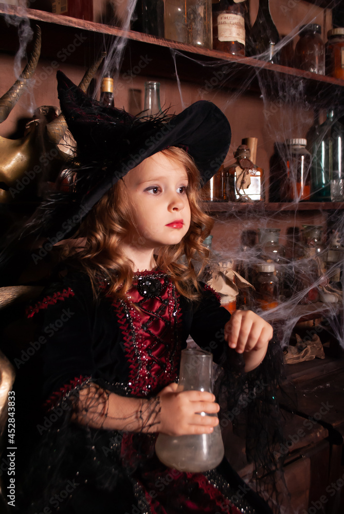 a child witch on Halloween with a flask in his hands looks into the distance in the web and smoke. Brunette girl in a witch costume a child witch on Halloween with a flask in his hands looks into the distance in the web and smoke. Brunette girl in a witch costume