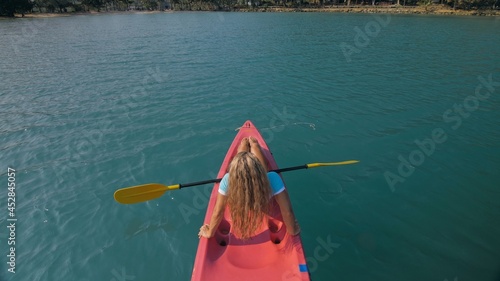 Seductive sexy blonde woman in blue swimsuit sails on pink plastic kayak along azure sea at exotic resort backside upper above view. Traveling to tropical countries. Girl is sailing on kayak in ocean.