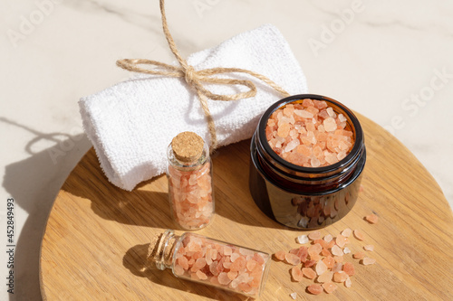 Pink Himalayan salt in the glass bottles with rolled towel on wooden board. Close up of pink crystals of sea salt. Spa body care concept