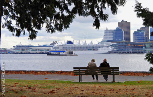 Couple romance sitting on park bench in Stanley Park, Vancouver Canada with picturesque city skyline view of downtown and cruiseships cruise ship liners at Canada Place terminal for Alaska cruising