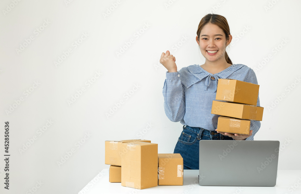 Young Asian woman business owner with many parcel boxes on the table happy online sales job, use the laptop, get an order from customers, take notes, and make arrangements for delivery by post Young Asian woman business owner with many parcel boxes on the table happy online sales job, use the laptop, get an order from customers, take notes, and make arrangements for delivery by post
