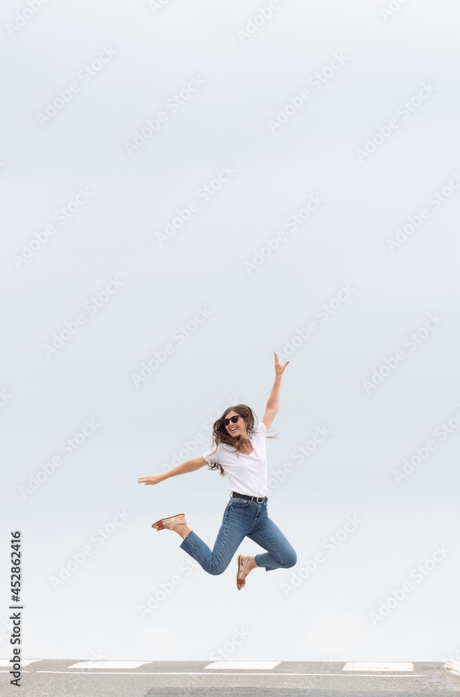 Happy casual dressed woman jumping on the zebra crossing of the road. Happiness concept with copy space. Minimal composition with asphalt and sky