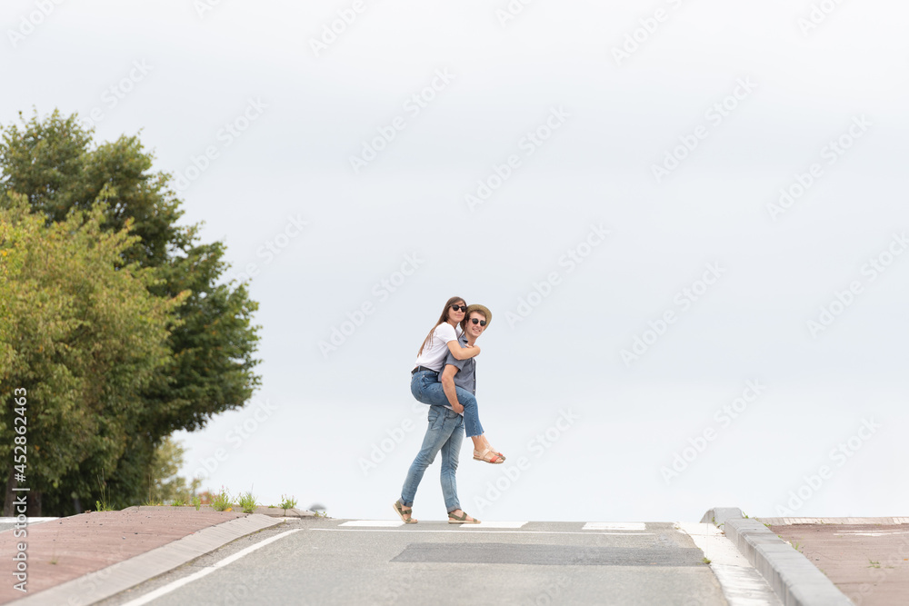 Romantic couple crossing the road. Young man carrying on his back or piggyback girlfriend. Minimal composition with copy space.