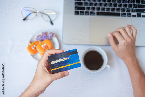 Fototapeta Naklejka Na Ścianę i Meble -  Hand holding credit card for online shopping on laptop during eating Halloween Cookies and coffee. Happy Halloween, Hello October, fall autumn, Festive, party and holiday concept