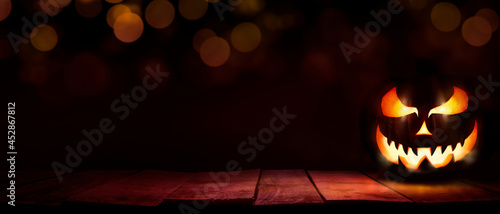 A scary halloween lantern with evil eyes and face on a rustic wood table with a spooky dark red background with faint light bokeh.